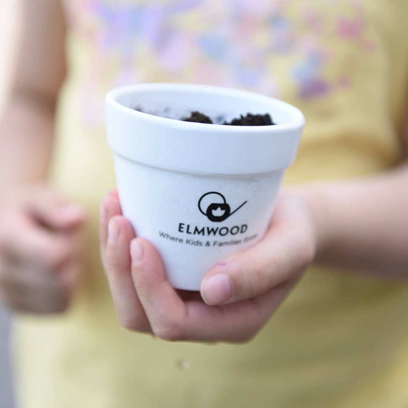 A child’s hands holding a small Elmwood pot of soil that reads “Where Kids & Families Grow”